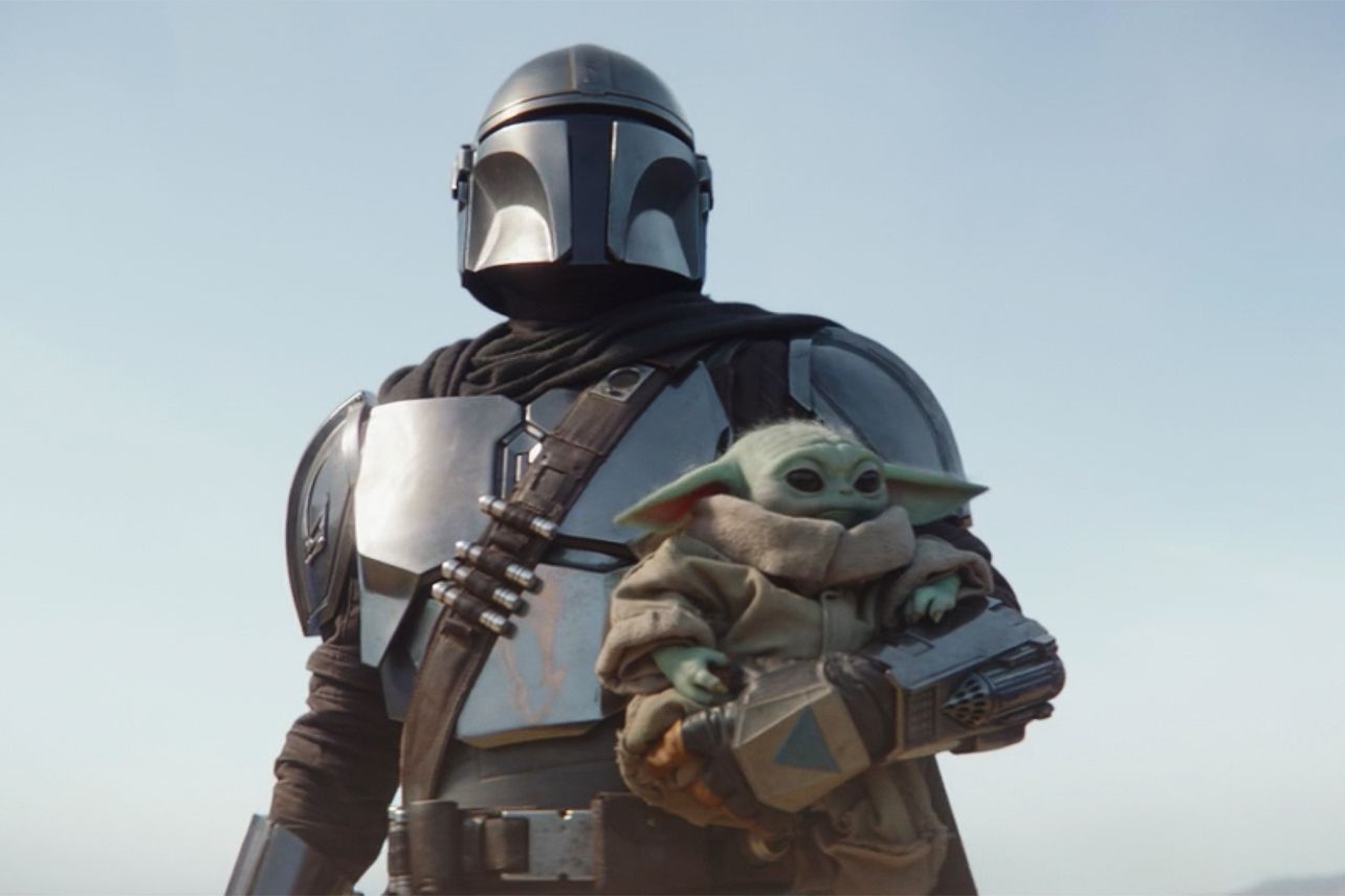 A still from TV show The Mandalorian showing main character Din Djarin holding Baby Yoda/The Child/Grogu.
