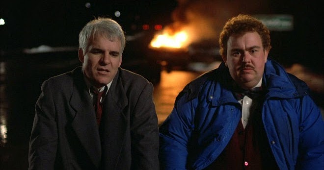 Neal Page (Steve Martin) and Del Griffith (John Candy) sit in the middle of the road as their car burns behind themin a still from "Planes, Trains and Automobiles."