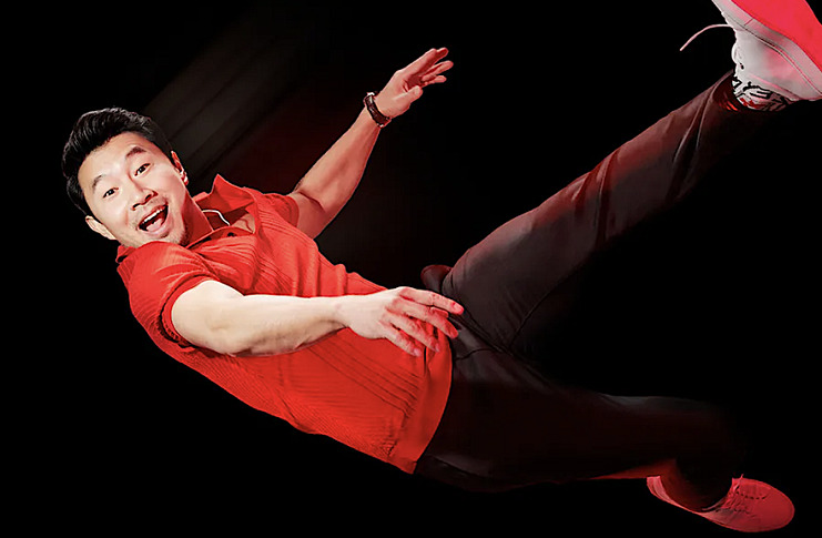 Simu Liu in a red shirt, black pants and red sneakers falling iwth a happy surpised look on his face.